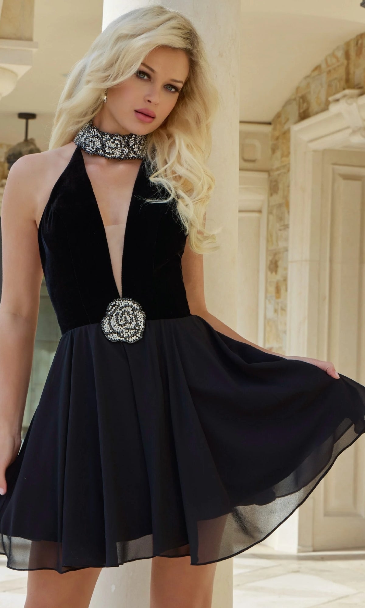 A woman with long blonde hair models the Jovani 45368 bead-choker short halter hoco dress—a sleeveless black cocktail dress with a deep V-neckline, beaded choker collar, and decorative brooch—outside a stone building.